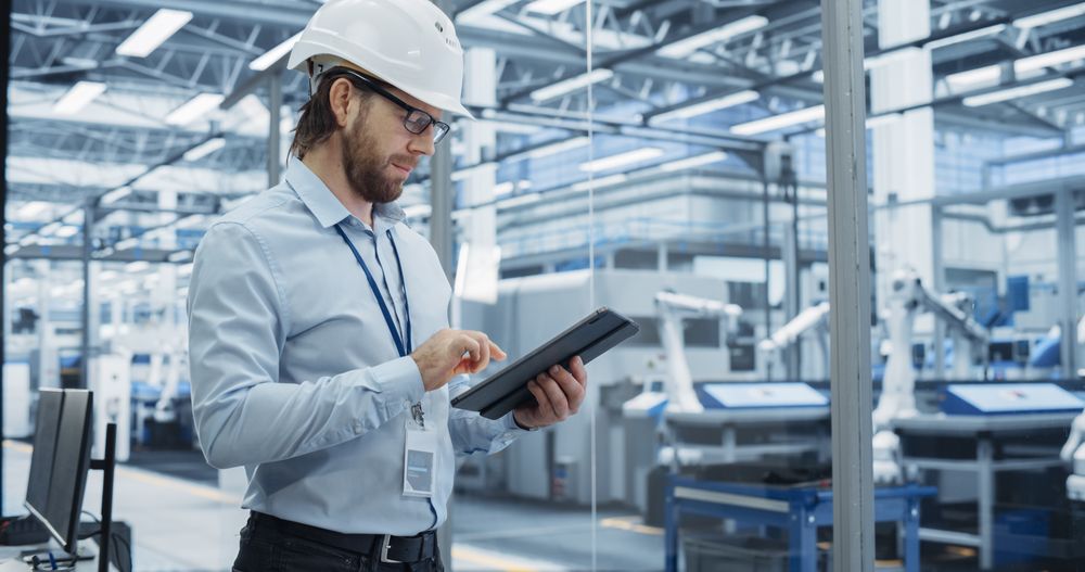 Electronics Factory Manager Working on Tablet at an Industrial Manufacture with Robotic Arms. Specialist in a Hard Hat Monitoring Big Data on a Computer, Checking and Planning Production Numbers