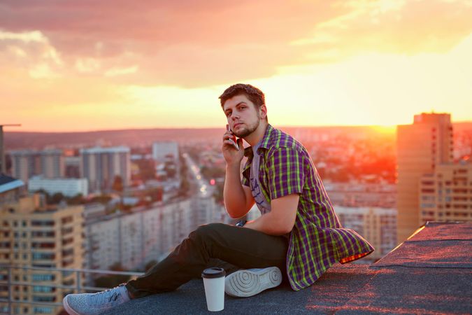 Male taking phone call while sitting on roof next to takeaway coffee