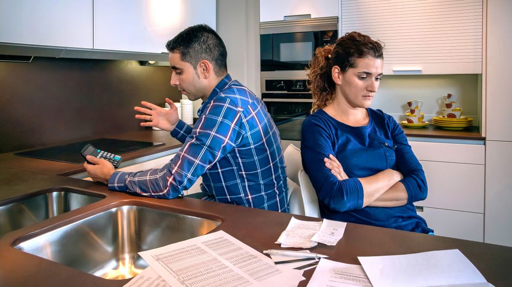 Angry couple with their backs to each other in kitchen with bills on counter