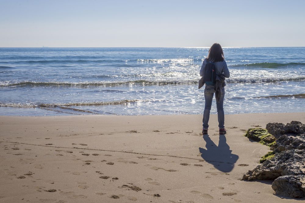 Back of female dressed in blue standing on the beach looking at the horizon