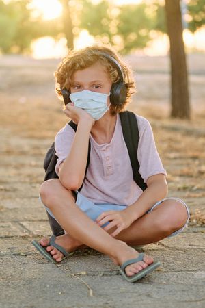 Boy in mask sitting on paved walkway and looking at camera