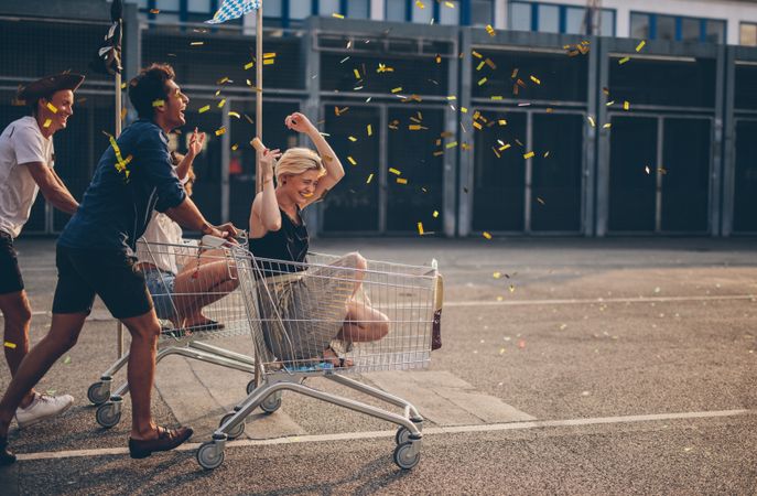 Multiethnic young people racing with shopping cart and blowing confetti