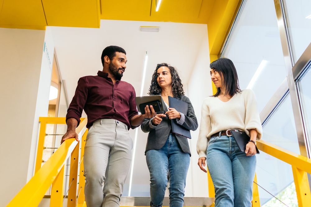 Multiracial coworkers happily talking about the company while walking down some yellow stairs in the office corridor