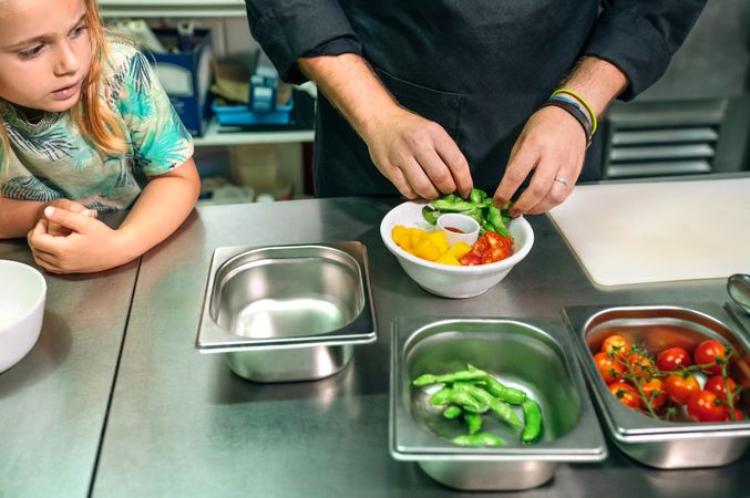 Unrecognizable chef preparing vegan poke bowl while little girl watching him in professional kitchen