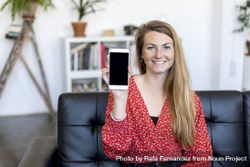Smiling woman showing a screen of a smart phone while sitting on a sofa 5ngB9Q