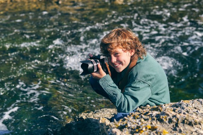 Smiling boy taking photo on rocky shore near stream of water