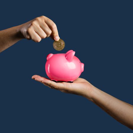 Hand inserting a Bitcoin coin into a pink piggy bank held by another hand on a blue background.
