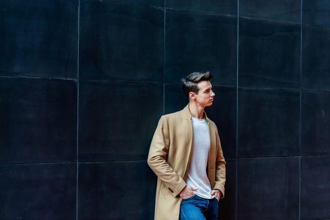 Young man leaning on dark wall outside looking away from camera dressed in camel coat 