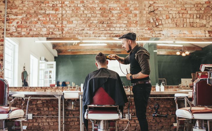 Hipster barber trimming customer’s hair in barbershop