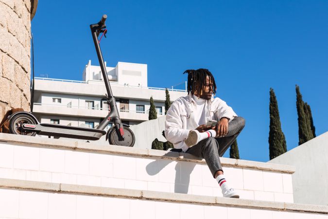 Black hipster man with scooter sitting on steps on mobile phone