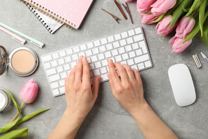 Woman types keyboard on grey background with tulips, top view