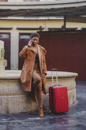 Stylish Black woman talking on smartphone with luggage on street