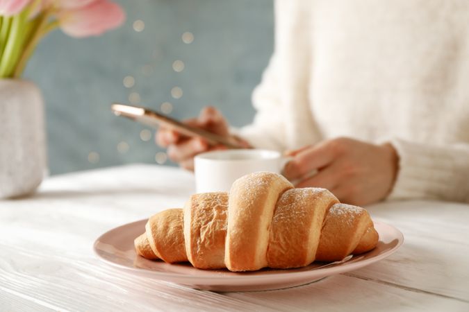 Person checking phone breakfast with croissant in foreground