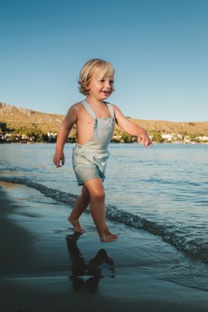 Barefoot positive little boy walking on sandy beach of wavy sea