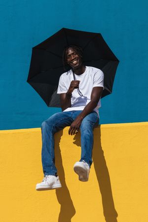 Happy Black man sitting on colorful wall with umbrella