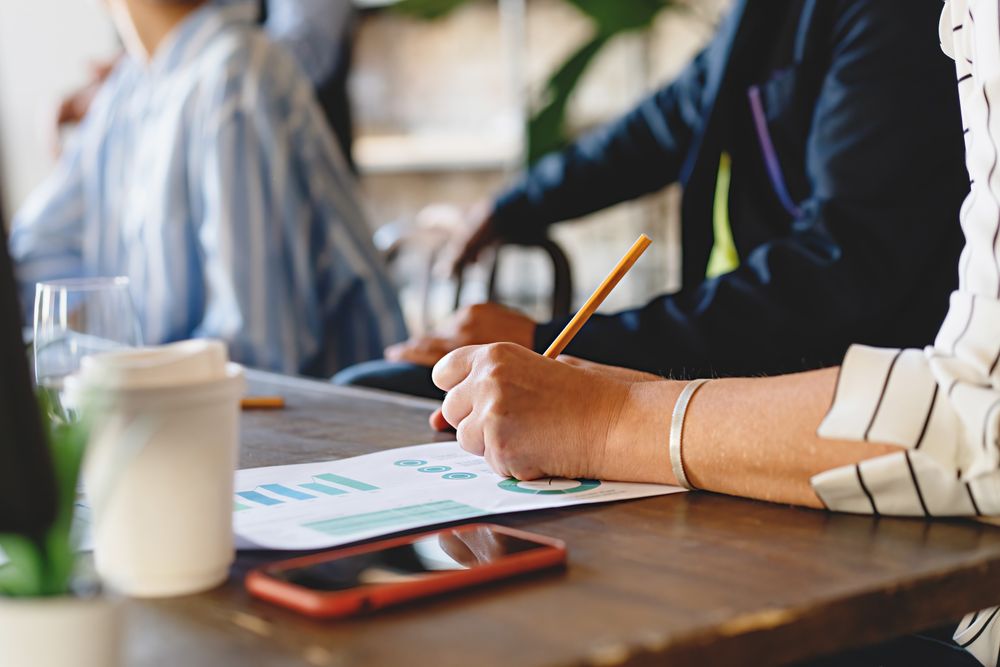 Close-up of woman's hand taking notes on chart paper in office meeting, wooden table with other people, papers, smartphone, and coffee cup