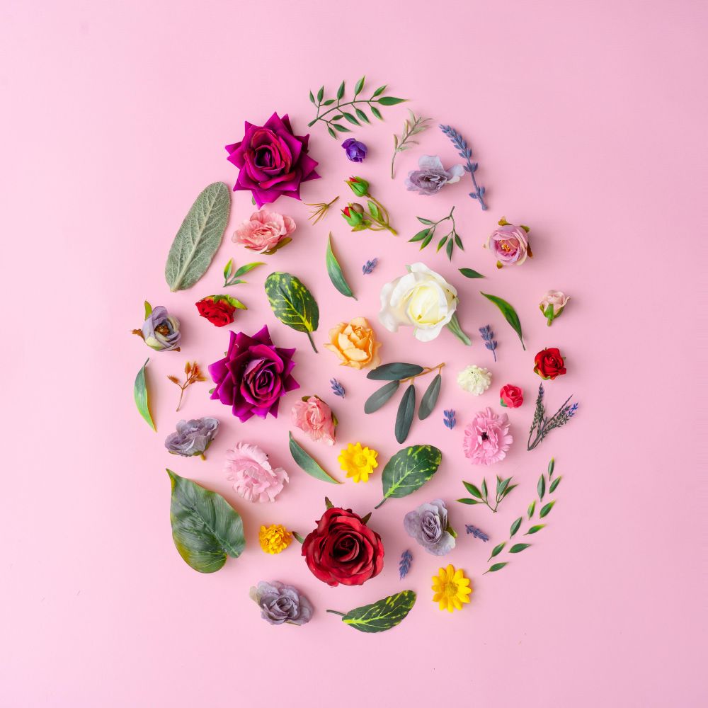 Easter egg shape made with various spring flowers and leaves on pastel pink background