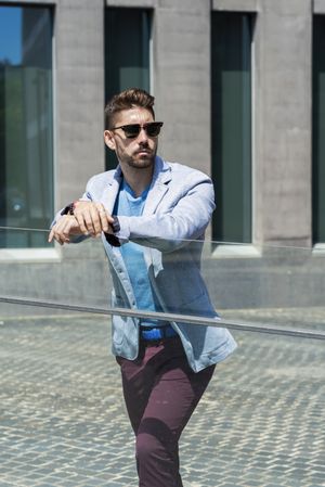 Portrait of a young bearded man with sunglasses leaning on glass railings outdoors in the city while looking aside