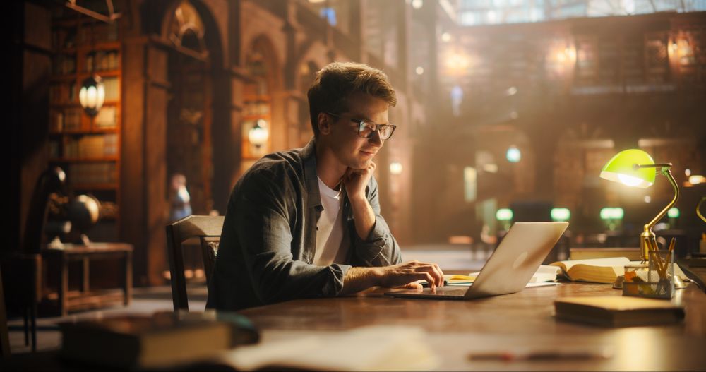 Focused Young Man Typing on Laptop in a Majestic Library, Illuminated by Soft