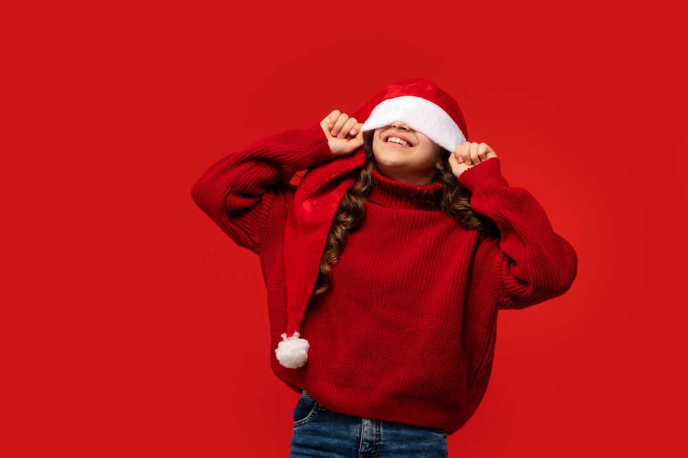 Smiling girl covering eyes with Santa hat in playful pose.