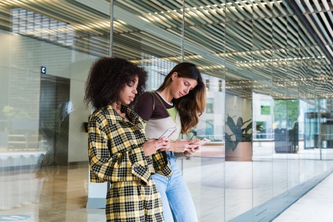 Multiracial girlfriends chatting on smartphones against glass wall