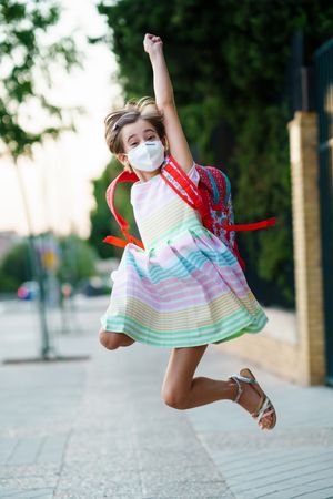 Happy girl jumping outside school gates wearing facemask