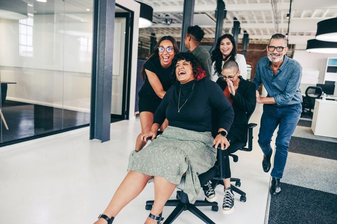 Carefree businesspeople having fun in a modern workplace pushing coworkers in office chairs