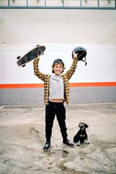 Cheerful child raising skateboard with helmet near dog on street