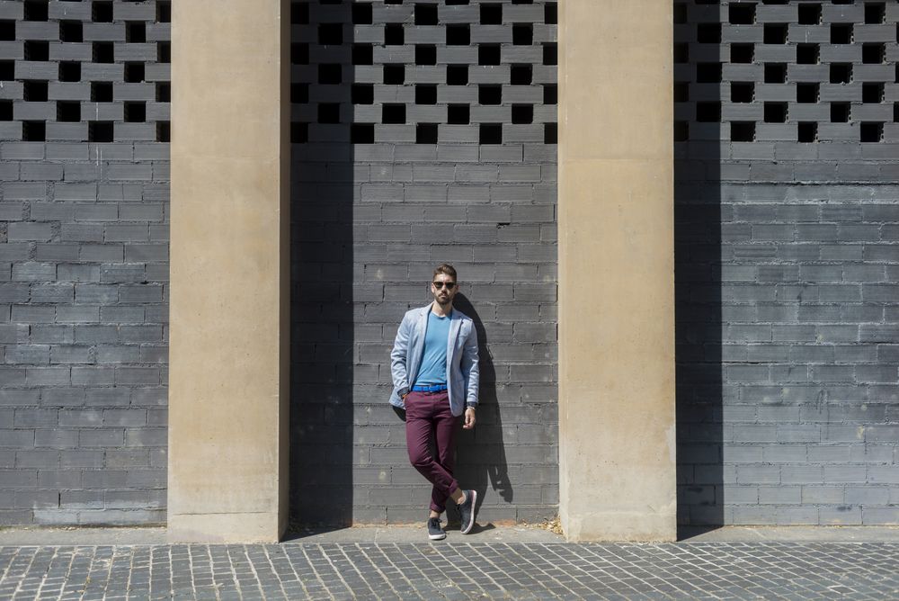 Young man in blazer leaning on a building wall outside looking at camera