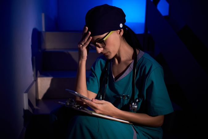 Worried woman in scrubs sitting on stairs and browsing smartphon