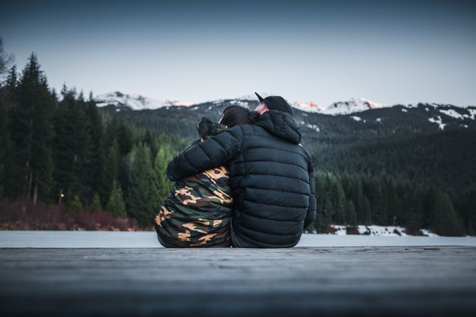 Two people hugging and sitting by lake near trees and mountains in view