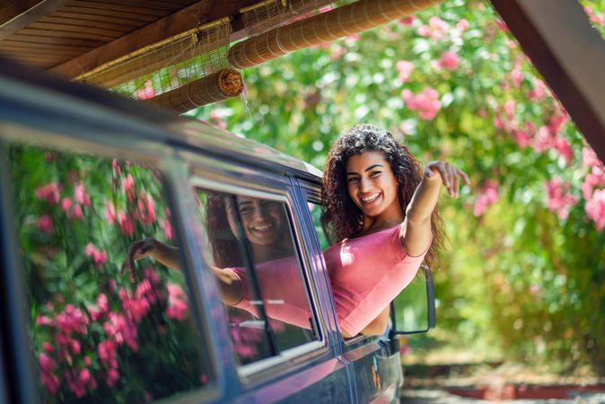 Happy female in pink t-shirt leaning out of car window with arm forward
