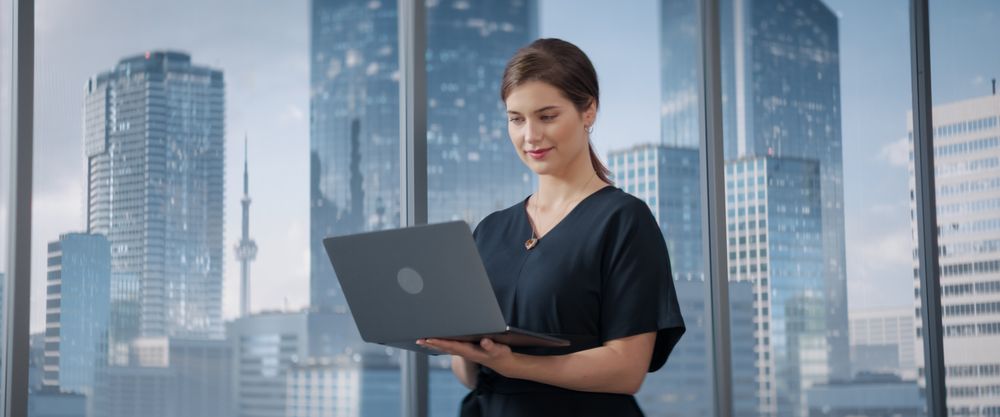 White Businesswoman Standing In Her Corner Office Next To Big Window With City View And Using Laptop Computer.