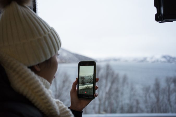 Calm woman recording video with smartphone of wintry scenery while looking out window