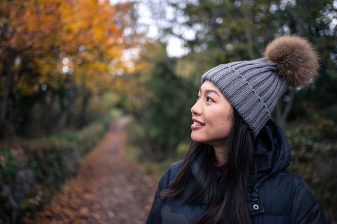 Smiling Asian woman in warm clothes standing in park with trees and stony fence