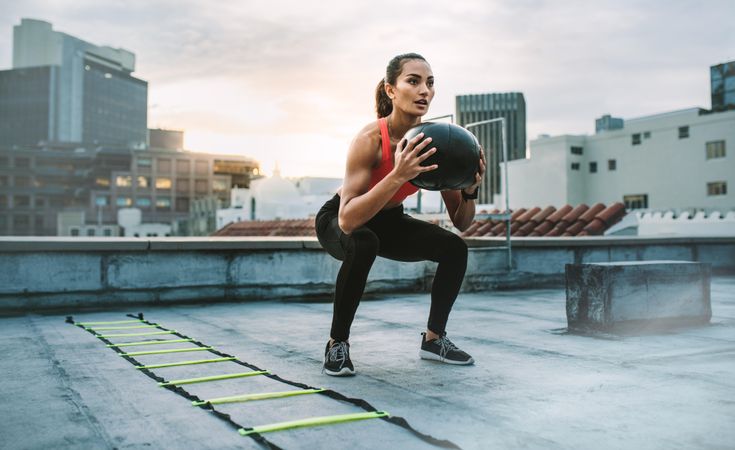 Female athlete doing squats holding a medicine ball standing on a rooftop