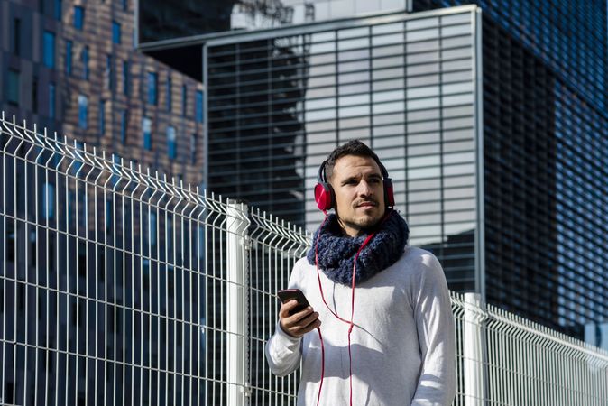 Young man walking in scarf past metallic fence and looking up from smartphone