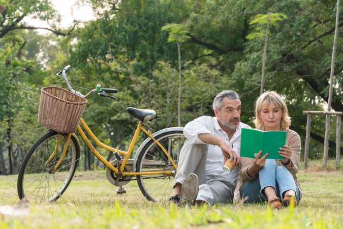 Happy older man and woman sitting on grass to reading book at park