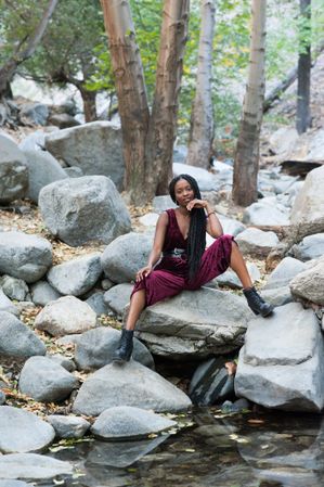 Fashionable Black woman sitting on boulders above a creek looking at camera
