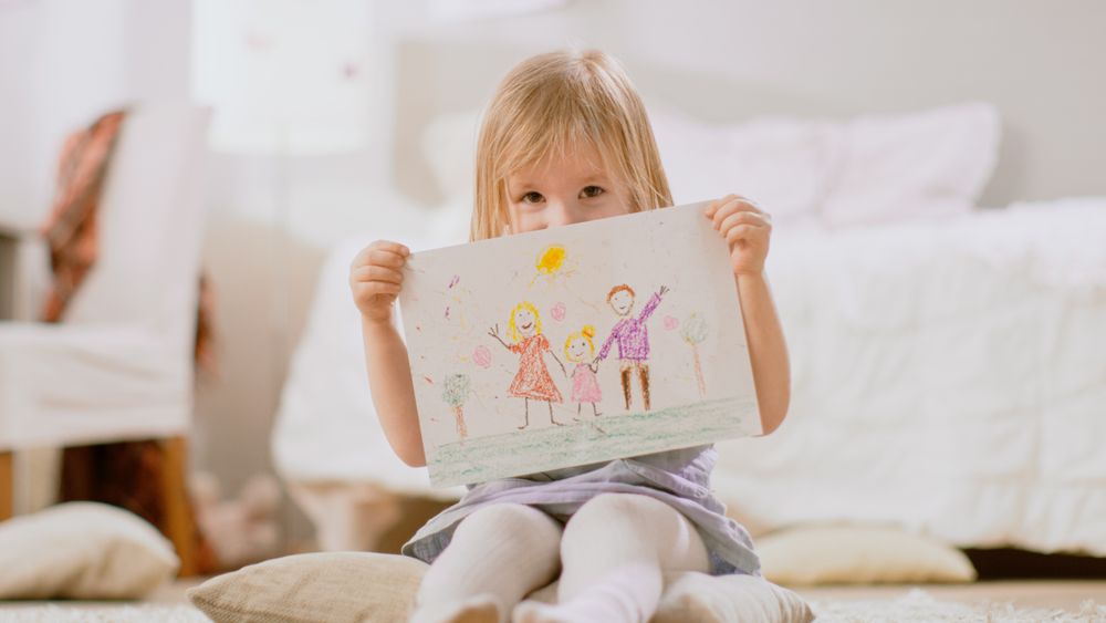 Young Girl Sitting on Pillows and Shows Drawing of Her Family and Hides Behind It.