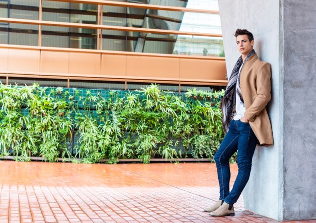 Serious man in camel coat leaning on wall outside