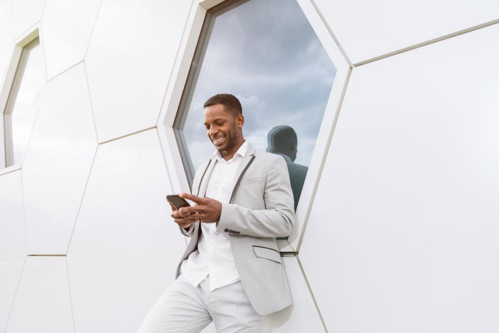 Elegant Black man browsing smartphone leaning on geometrical wall