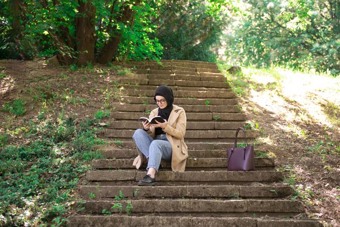 Woman in headscarf sitting on outdoor stairs immersed in a book