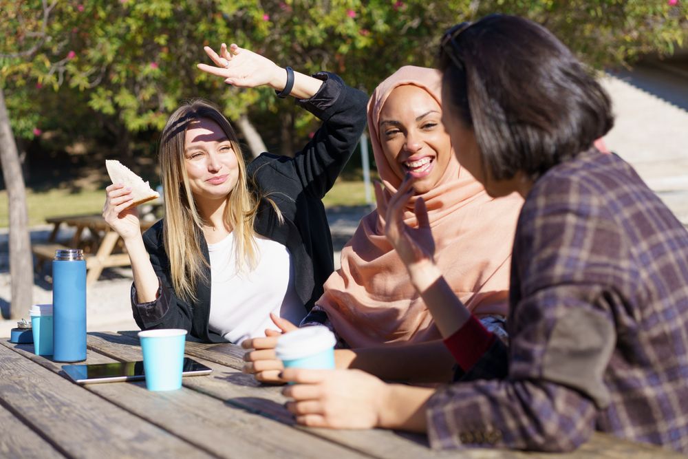 Three women laughing outside on park bench with insulated mugs and sandwiches
