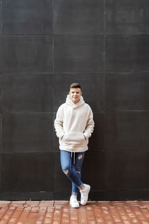 Young man in bright hoodie leaning on dark wall outside, vertical composition
