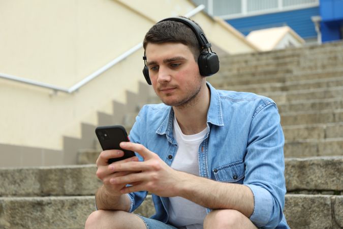 Young man in headphones sits on the steps and looking in phone