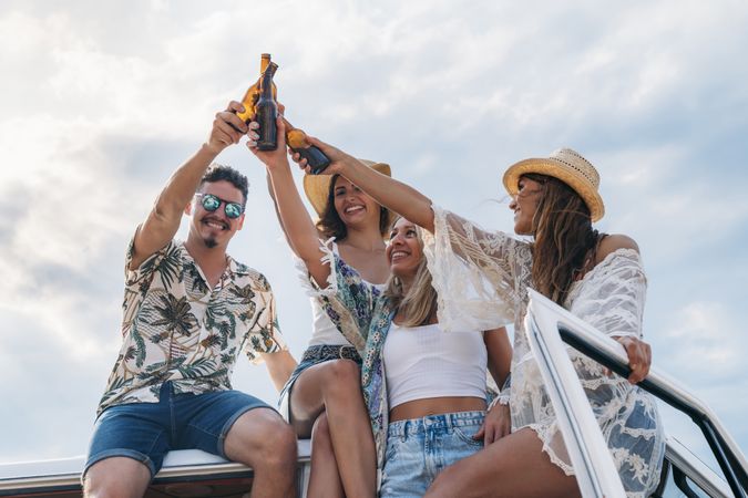 Smiling friends clinking bottles celebrating vacation on car roof in bright day