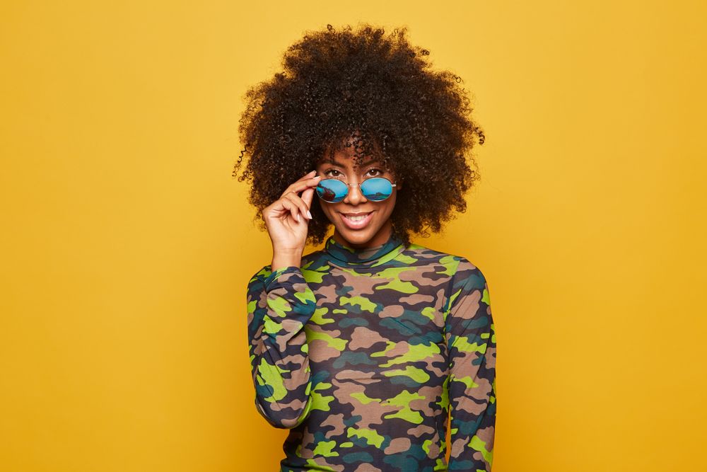 Trendy Black woman in sunglasses on bright backdrop