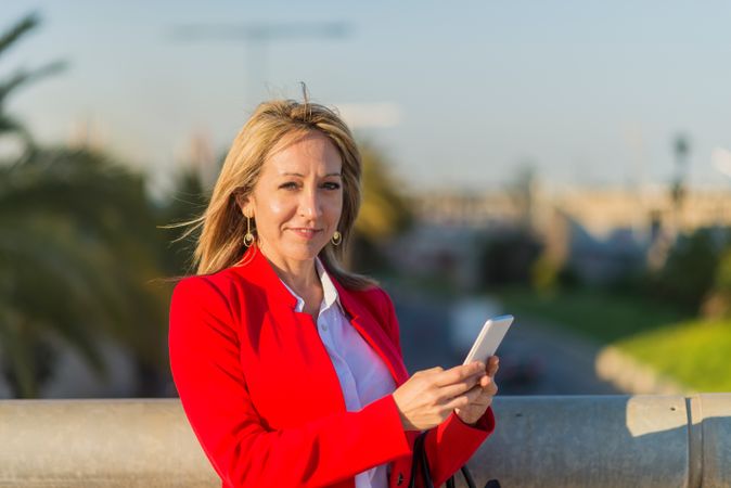 Smiling blonde businesswoman wearing red jacket using a phone standing on the street