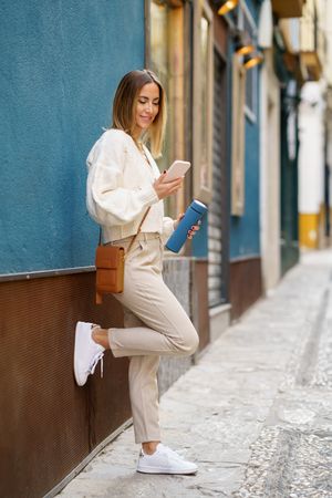 Happy woman in cream leaning on wall outside and checking phone